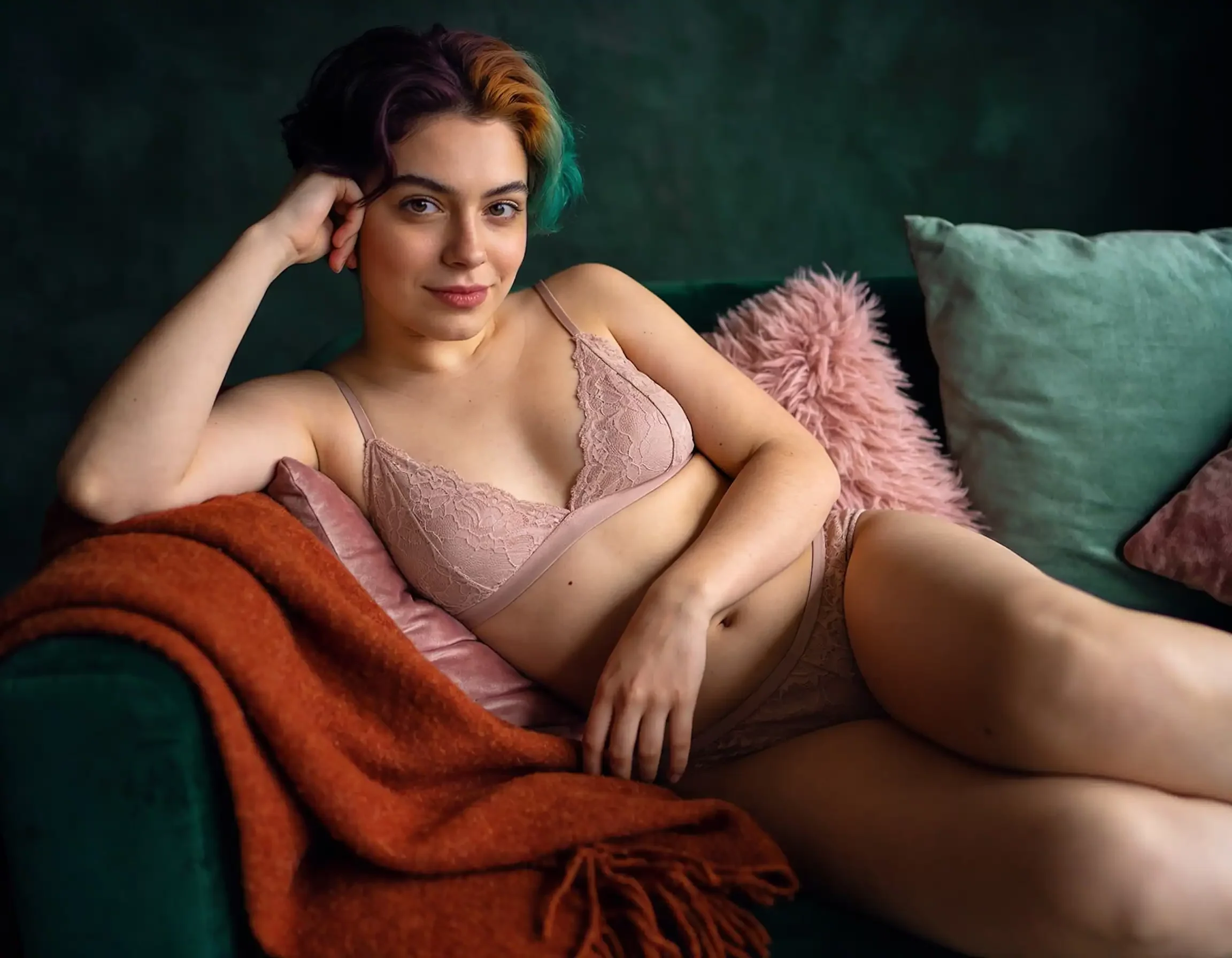 Woman wearing pink lace lingerie reclining on a green couch with orange blanket, posing for a boudoir-style shot, multicolor hair visible.